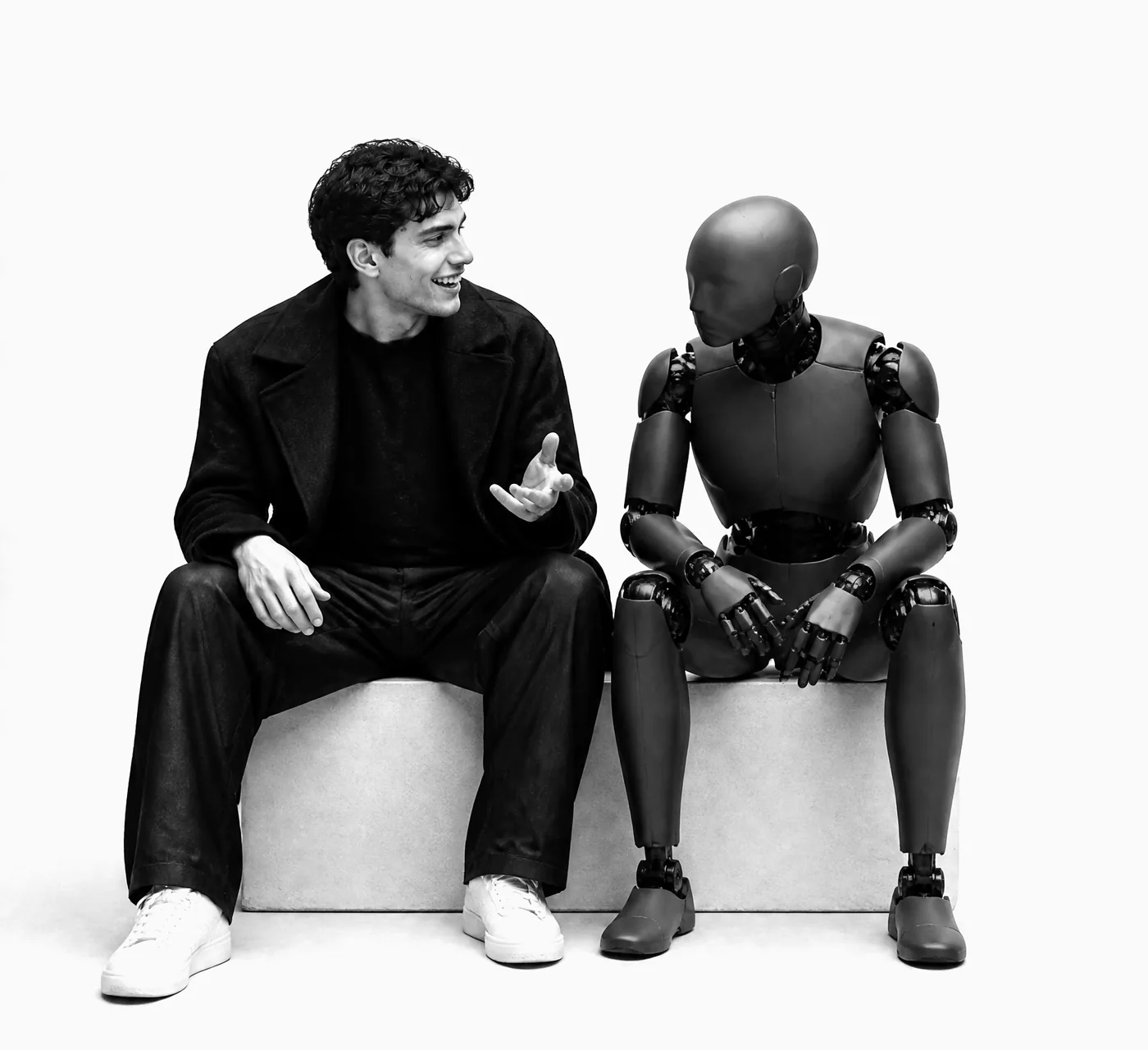 A young man in a dark coat sits on a plinth beside a robot, laughing and gesturing mid-conversation while the robot leans in to listen.