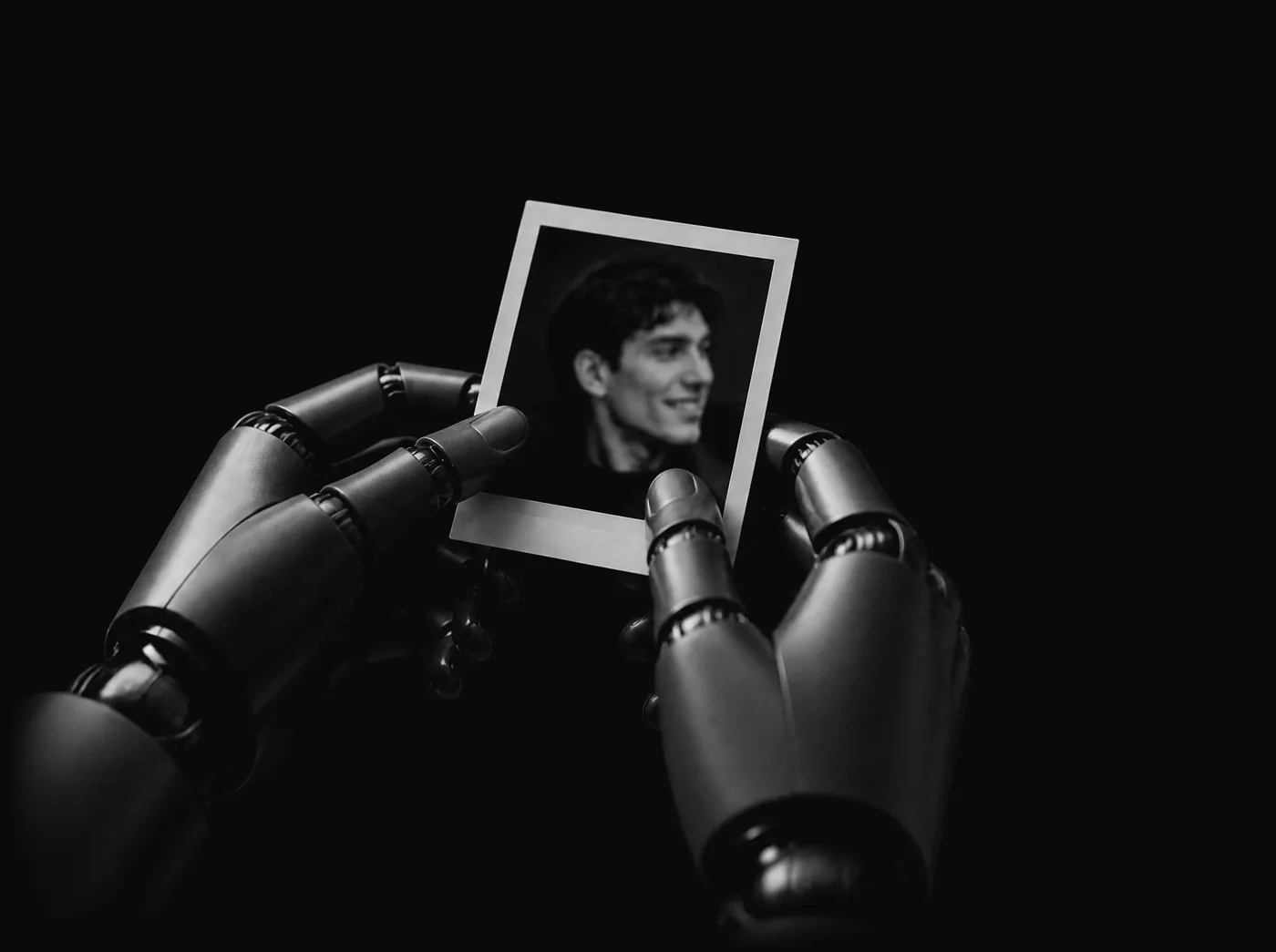 Two robot hands carefully hold a small instant photograph of a smiling young man.