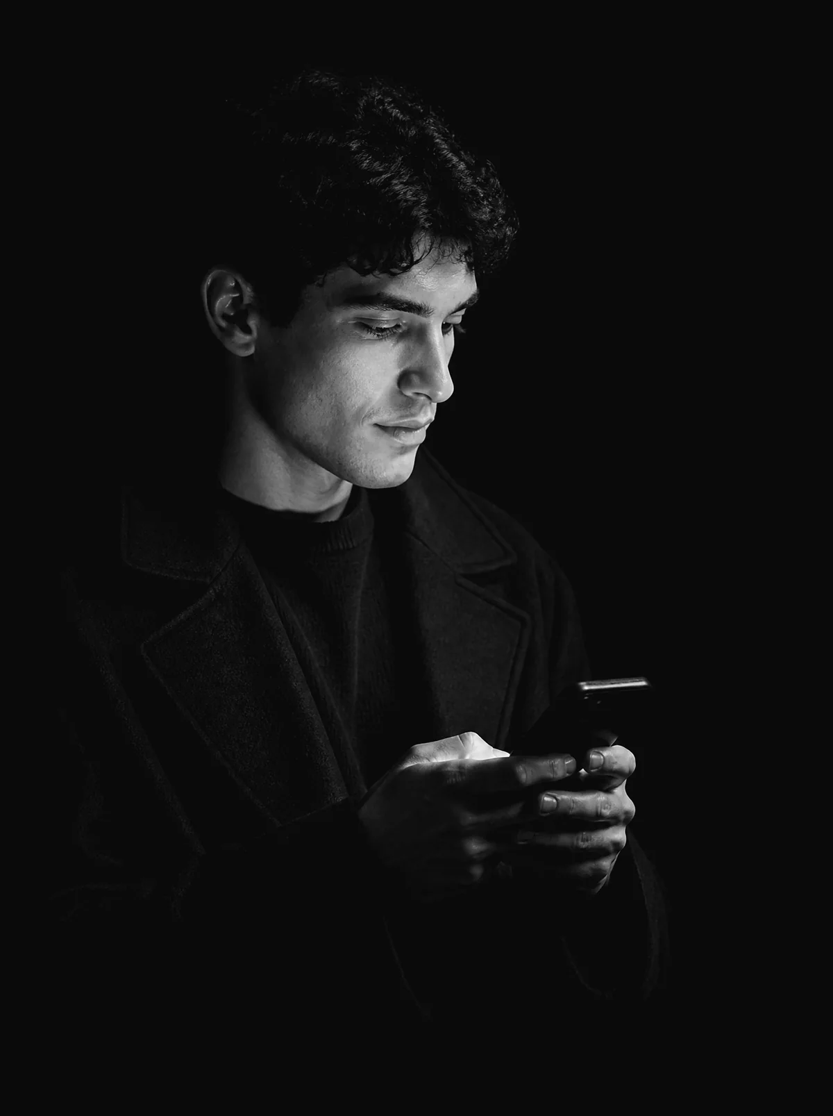 A young man in a dark coat looks down at his phone with a small smile, his face lit by the screen.